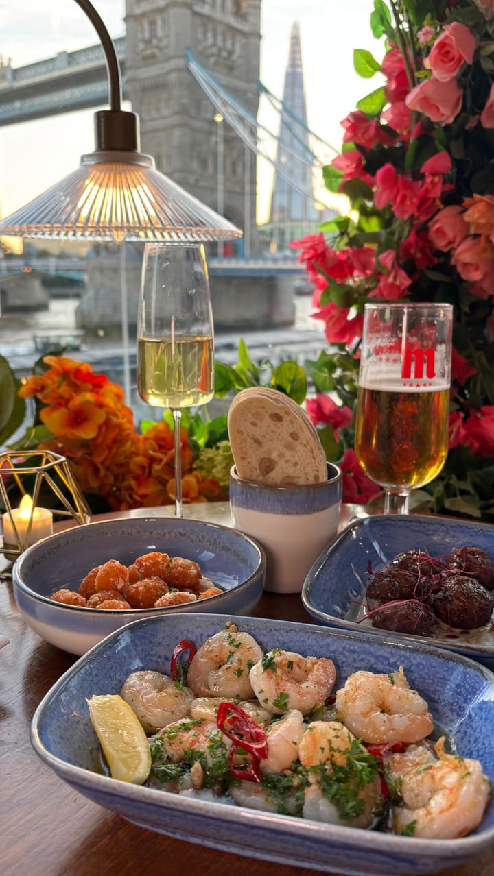 The video showcases a dinner experience at 'Vu from The Tower,' a restaurant with prominent views of London's Tower Bridge. It begins with the sunset view, then moves inside to show the menu, drinks (champagne and beer), and a variety of dishes. The food featured includes starters like lamb koftas, tiger prawns, and squid balls. The main courses shown are a sirloin steak with chips and a penne alla vodka with smoked salmon and caviar. The meal concludes with a chocolate and raspberry tart for dessert, followed by a final shot of the illuminated Tower Bridge at night.