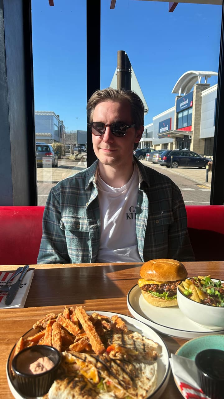 A couple visits a TGI Fridays to showcase their 'Everyday Trio' value menu, featuring shots of the menu, the interior, and various dishes including garlic bread, loaded chicken bites, a chicken burger, quesadillas, and fries.