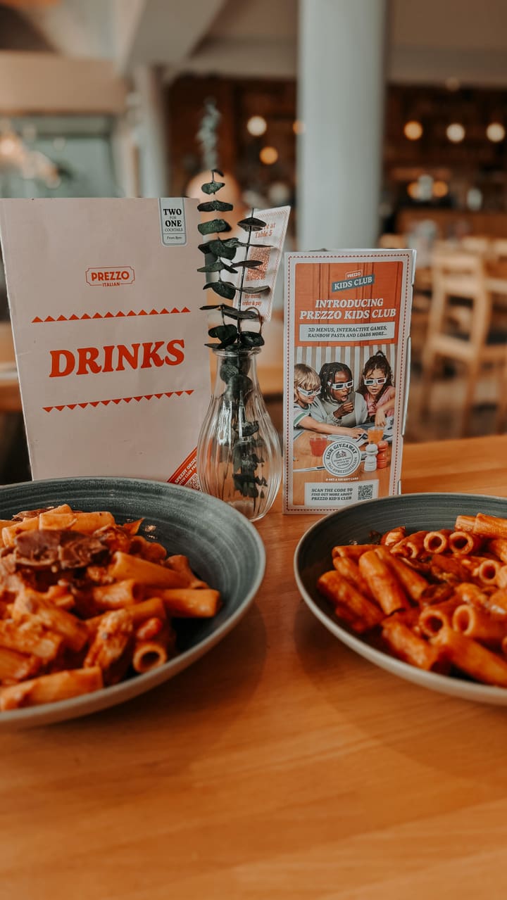 The video promotes the Italian restaurant chain Prezzo, specifically highlighting its new 'Kids Club'. It begins with an exterior shot of the restaurant, then moves inside to show the table setting and a young girl coloring with the kids' activity pack. The video showcases several dishes, including a chicken schnitzel with spaghetti and two different rigatoni pasta dishes. A key shot features parmesan cheese being freshly grated over the schnitzel dish. The video concludes with the young girl happily eating.