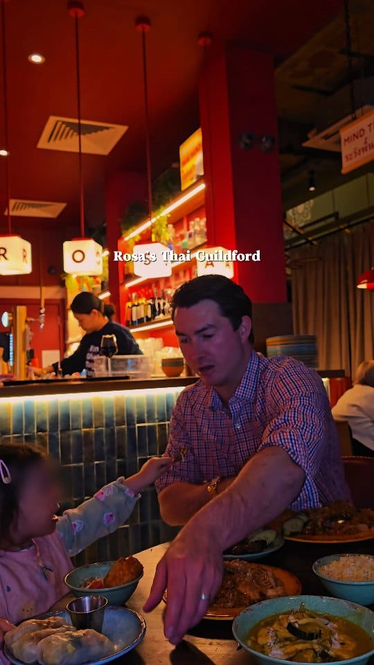 The video showcases a Thai restaurant experience at Rosa's Thai Guildford. It features various Thai dishes including fried rice, skewers, spring rolls, and fresh spring rolls with dipping sauce. It also shows a drink in a mason jar with a straw, and a toast with a beer glass. The video includes shots of the restaurant exterior, interior decor (neon sign, bar area), and the influencer and her child enjoying the meal. Text overlays identify the restaurant and mention a 

20 per person set menu.