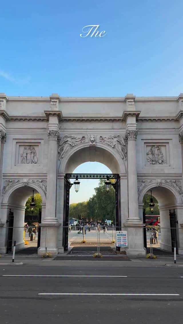 The video is a promotional montage for The Marble Arch Hotel in London. It begins with an exterior shot of the Marble Arch monument, followed by shots of the hotel's interior, including the lobby with floral arrangements, a hotel room with a seating area and bed, and a welcome platter with sparkling rosé, desserts, and snacks. It then showcases the hotel's restaurant, gym with a treadmill and elliptical machine, and an on-site cafe/shop with pastries and drinks. The video concludes with an exterior shot of the hotel's flag and sign.