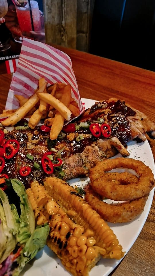 A promotional video for TGI Fridays, celebrating the 4th of July. It showcases the restaurant's festive decorations, including red, white, and blue balloons, American flags, and a 'Happy 4th of July' sign. The video features a meal, starting with appetizers: sesame chicken strips, mozzarella planks, and loaded potato skins. The main courses shown are a rump steak with fries, onion rings, and corn on the cob, and a large rack of BBQ ribs. The meal concludes with a chocolate fudge cake with ice cream for dessert. The video includes close-up shots of the food being eaten, such as dipping a potato skin and pulling apart a mozzarella stick.