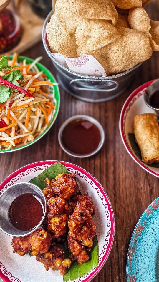 The video begins with an exterior shot of Rosa's Thai restaurant, showing the red facade and outdoor seating. The influencer then enters the restaurant. Inside, there are shots of the interior, including tables, decor, and the kitchen area. Close-ups of various Thai dishes are shown, including papaya salad, spring rolls, sweetcorn fritters, chilli and basil chicken with rice and cucumber, and green curry with rice. There are also shots of drinks being served and the influencer sitting at a table. The video ends with a shot of a banner detailing 'Rosa's Story' and 'Chef Saiphin'.